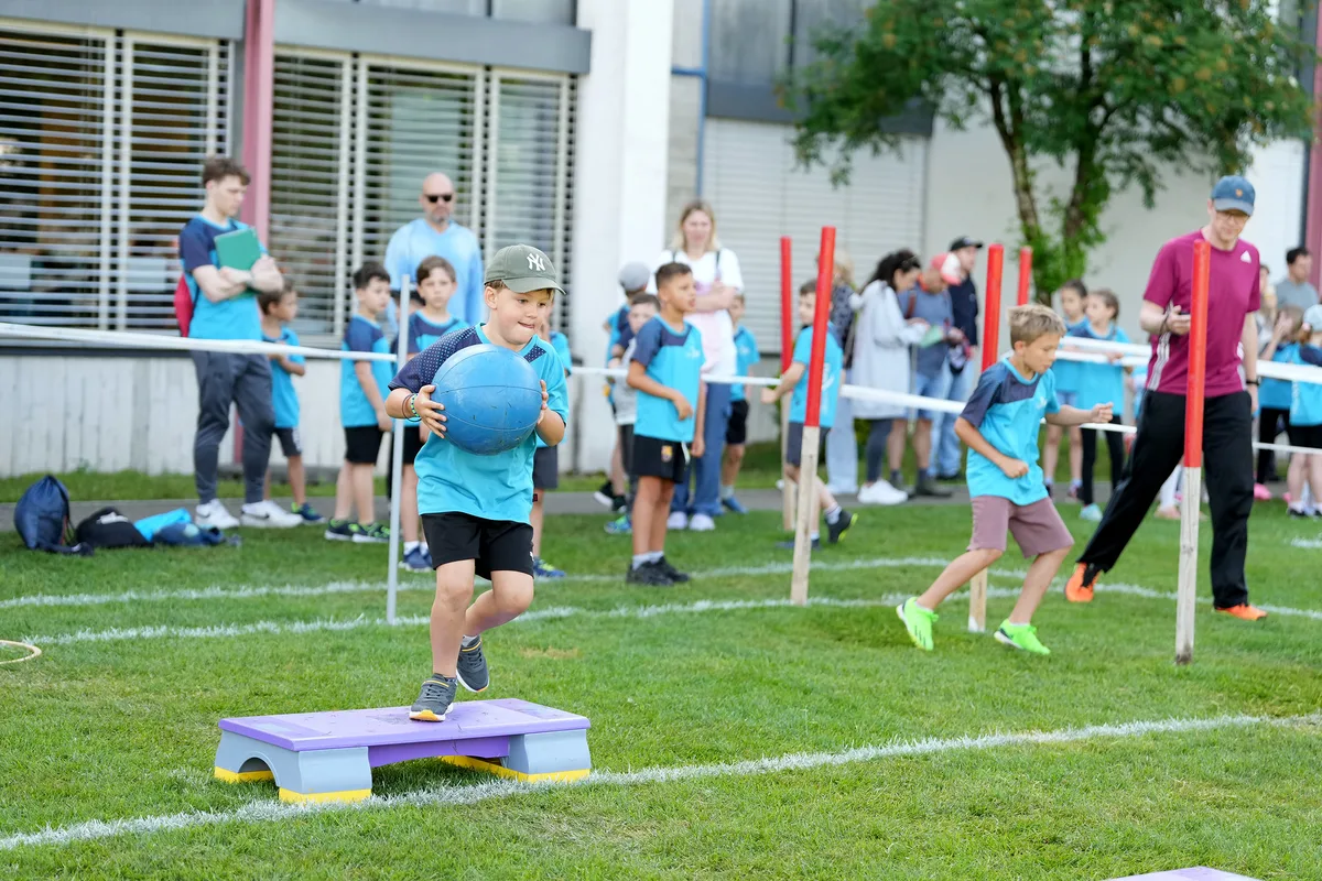 Kinder auf einem Turnparcours. Eines hält einen blauen Ball.